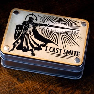 May include: A metal box with a wood top featuring a black and white illustration of a knight wielding a sword. The text "I cast smite" is printed on the box.