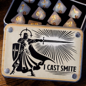Può includere: Una scatola di legno con un'illustrazione in bianco e nero di un cavaliere che tiene una spada e il testo "I cast smite". La scatola è aperta e contiene un set di dadi bianchi con numeri dorati.