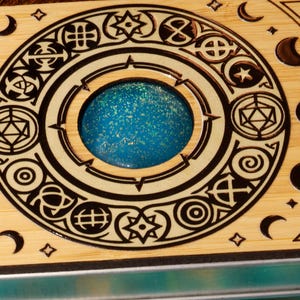 K&ouml;nnte beinhalten: Eine Holzkiste mit einem kreisf&ouml;rmigen Design, das ein mit blauem und wei&szlig;em Glitzer gef&uuml;lltes Zentrum aufweist. Die Kiste hat einen schwarzen Rand und ist mit verschiedenen Symbolen und Mondsicheln verziert.
