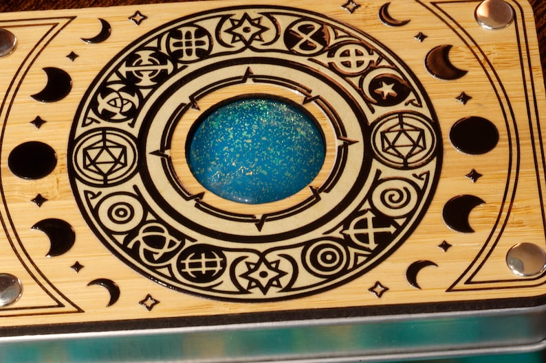 K&ouml;nnte beinhalten: Eine Holzkiste mit einem kreisf&ouml;rmigen Design, das ein mit blauem und wei&szlig;em Glitzer gef&uuml;lltes Zentrum aufweist. Die Kiste hat einen schwarzen Rand und ist mit verschiedenen Symbolen und Mondsicheln verziert.