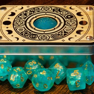 K&ouml;nnte beinhalten: Eine Holzkiste mit einem Metalldeckel und einem dekorativen Design eines Kreises mit Symbolen und Monden. Die Kiste ist ge&ouml;ffnet und enth&auml;lt einen Satz tealblauer W&uuml;rfel mit goldenen Zahlen.
