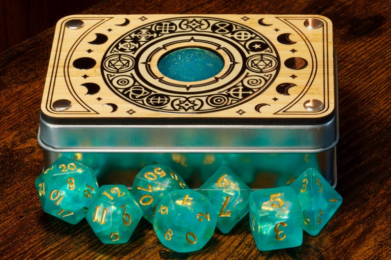 K&ouml;nnte beinhalten: Eine Holzkiste mit einem Metalldeckel und einem dekorativen Design eines Kreises mit Symbolen und Monden. Die Kiste ist ge&ouml;ffnet und enth&auml;lt einen Satz tealblauer W&uuml;rfel mit goldenen Zahlen.