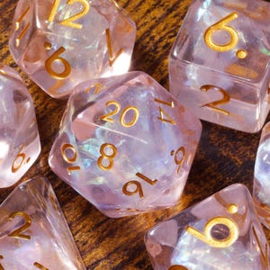 May include: A set of seven polyhedral dice made of translucent, pale pink resin with an iridescent, pearlescent shimmer inside. Each die features engraved numbers filled with gold-coloured paint, including a twenty-sided die showing the number 20.