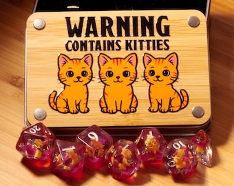 Könnte beinhalten: Eine Holzbox mit dem Text "WARNING CONTAINS KITTIES" und drei orangefarbenen Cartoon-Katzen. Darunter befinden sich mehrere rote und transparente Würfel mit Katzenfiguren. Die Würfel sind für Tischspiele.