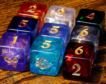 May include: A collection of eight translucent, multi-colored six-sided dice arranged in a grid. Each die features gold-colored numerals on its faces and contains iridescent, reflective inclusions suspended within the clear, colored resin material.