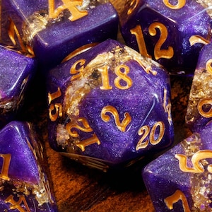 May include: A set of polyhedral dice in a deep purple resin with a shimmering, glittery finish. Each die features embedded gold-colored foil flakes and engraved numbers filled with gold paint, creating a distinct contrast against the purple.