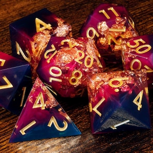 May include: A set of seven polyhedral dice featuring a deep purple and blue gradient resin finish with embedded gold-coloured metallic flakes. Each die has engraved numbers filled with gold paint, including a d20, d12, d10, d8, d6, d4, and a d100.