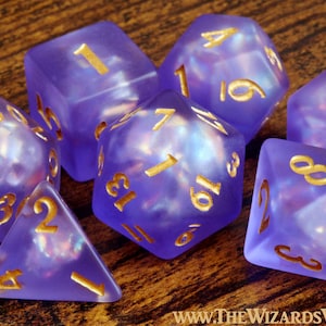 Peut inclure: Un ensemble de sept dés polyédriques violets translucides avec des chiffres dorés. Les dés présentent un effet intérieur nacré et nuageux. Le texte "WWW.THEWIZARDSVAULT.COM" est imprimé en petites lettres blanches en bas à droite.