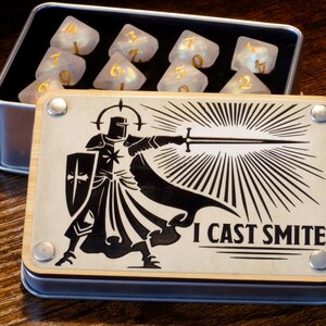 May include: A wooden box with a black and white illustration of a knight holding a sword. The box has the text "I cast smite" printed on it. The box is open and contains a set of white dice with gold numbers.