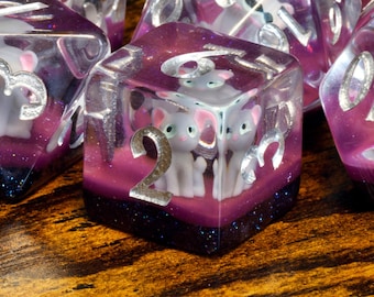 May include: Close-up of a purple and silver dice with a glitter finish. The dice has a number "2" on one side and a picture of three white kittens on the other side.