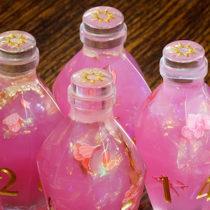 May include: Four pink glass bottles with gold accents and clear lids. Each bottle has a different number on the front, 1, 2, 14. The bottles are filled with a pink liquid and have small pink flowers inside.