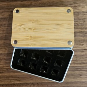 a wooden board with a black rectangular object inside a metal container on a wooden surface.