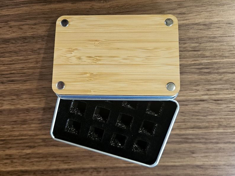 a wooden board with a black rectangular object inside a metal container on a wooden surface.