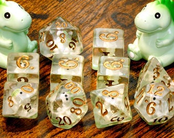 May include: A set of polyhedral dice made of clear resin, each containing a small white dinosaur figure inside. The numbers on the dice are engraved in gold. Two larger, light green dinosaur figurines are positioned on either side of the dice.