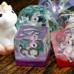 May include: A set of six clear dice with unicorn figures inside. The dice are in various colors, including purple, green, pink, and yellow. The dice are on a wooden surface next to a white unicorn figurine.