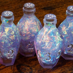 May include: Four iridescent, faceted dice with a bottle-like shape. The dice are primarily purple with internal rainbow-coloured reflections. Each die has a number etched on its surface, and a star design on the top. The dice are on a wooden surface.