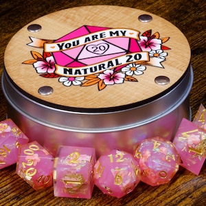 May include: A pink and white dice set with gold glitter and floral accents in a tin box with a wood lid. The lid has a pink and white dice with the text "You are my natural 20".