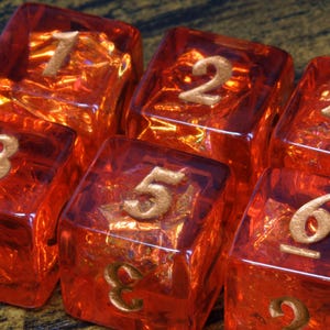 May include: A set of six translucent orange cubic dice with rounded edges. Each die features gold-coloured engraved numbers on the top faces, displaying the digits one through six in a sequential arrangement across the individual dice surfaces.