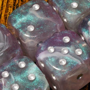 May include: A set of six-sided dice featuring a translucent resin construction with swirling purple and white clouds, embedded with fine teal glitter particles. Each die has rounded corners and white circular pips indented into the surface.