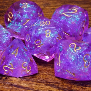 May include: A set of seven polyhedral dice made of translucent purple resin with embedded iridescent blue foil flakes and gold-coloured numbers. The collection includes a d20, d12, d10, d8, d6, d4, and a percentile die, all featuring sharp edges.