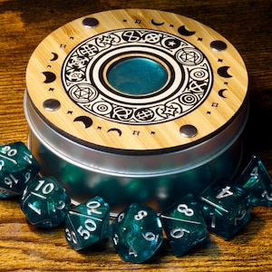 May include: A wooden box with a circular lid featuring a decorative design of moons, stars, and other symbols. The box is open and contains several teal-colored dice.