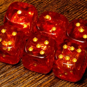 May include: A collection of six translucent red dice with rounded corners and gold-coloured pips. Each die features a distinct arrangement of pips on its visible faces, reflecting light through the deep red material to create a glowing effect.