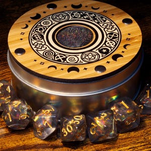 May include: A wooden box with a circular design featuring a black and white pattern of moons, stars, and geometric shapes. The box has a clear lid with a glittery surface. The box is surrounded by iridescent dice with gold numbers.