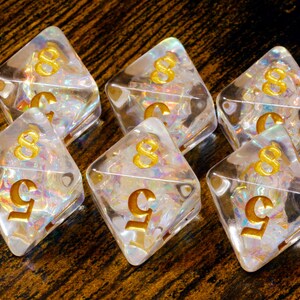 May include: Six clear, diamond-shaped dice with gold numbers. The dice have a shimmering, iridescent interior. The numbers are a stylized '8' and '5'. The dice are arranged on a dark, textured surface.