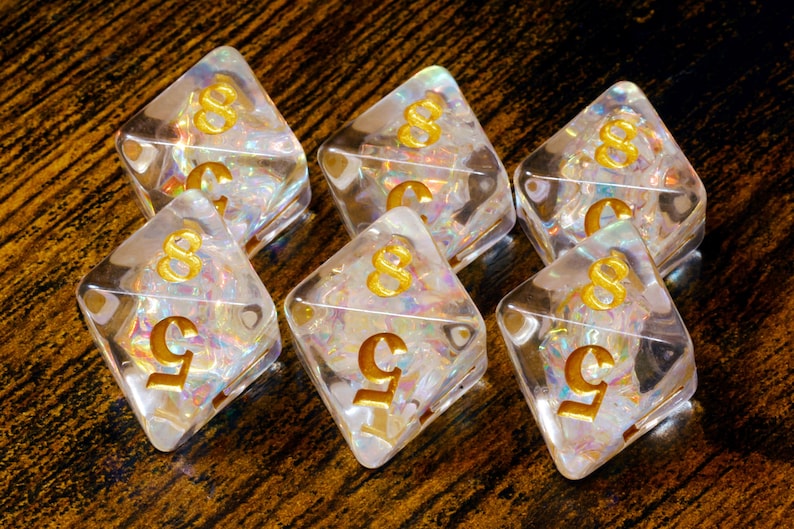 May include: Six clear, diamond-shaped dice with gold numbers. The dice have a shimmering, iridescent interior. The numbers are a stylized '8' and '5'. The dice are arranged on a dark, textured surface.