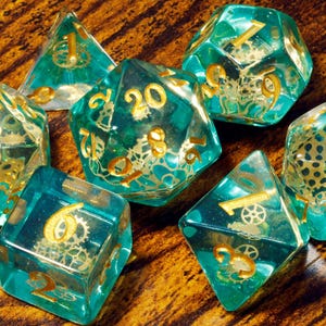 May include: A set of seven translucent teal polyhedral dice with gold-colored numbers and embedded metallic gear components visible inside each die. The set includes a d20, d12, d10, d8, d6, d4, and a percentile die, all featuring sharp edges.