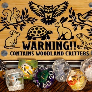 May include: A set of six clear dice with woodland animal illustrations inside. The dice are in a metal tin with a wooden lid that says "WARNING!! CONTAINS WOODLAND CRITTERS".