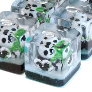 May include: A row of clear resin dice, each containing a Dalmatian dog design. The dice have a silver loop and a base with black, white, and green elements. The dice are cube-shaped.