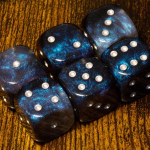 May include: A set of six-sided dice featuring a deep blue and black swirling galaxy-like pattern with glittery accents. Each die has rounded corners and white circular pips indicating the numerical values on each of the six faces of the cubes.