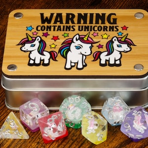 Op de afbeelding: Een metalen doos met een houten deksel waarop "Warning Contains Unicorns" staat. Het deksel heeft drie cartoon-eenhoorns met regenboogmanen en -staarten. De doos bevat acht transparante dobbelstenen met eenhoornbeelden erin.