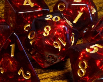 May include: A collection of translucent deep red polyhedral dice with gold-coloured engraved numbers. The set includes various shapes such as a twenty-sided die, a four-sided die, and other multi-faceted gaming dice with clear, legible numerals.