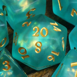 K&ouml;nnte beinhalten: Ein Satz tealblauer W&uuml;rfel mit goldenen Zahlen. Die W&uuml;rfel sind durchscheinend und haben eine schimmernde Oberfl&auml;che. Die Zahlen auf den W&uuml;rfeln sind 20, 8, 10, 16, 14, 7, 3, 2, 1, 5, 0 und 6.