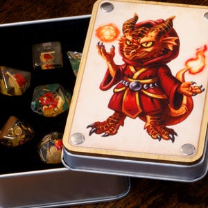 Op de afbeelding: Een metalen doos met een scharnierend deksel, met meerdere dobbelstenen. Het deksel toont een cartoon drakenpersonage in rode gewaden, met een gloeiende bol. De dobbelstenen zijn in verschillende kleuren en ontwerpen, geschikt voor tabletop games.