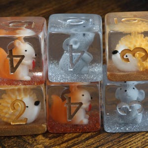 Könnte beinhalten: Sechs transparente Harzwürfel mit Miniatur-Tierfiguren im Inneren. Das Set enthält orangefarbene Eichhörnchen, graue Mäuse und gelbe Igel. Jeder Würfel hat Glitzer am Boden und große, goldene oder silberne metallische Zahlen auf der Vorderseite.