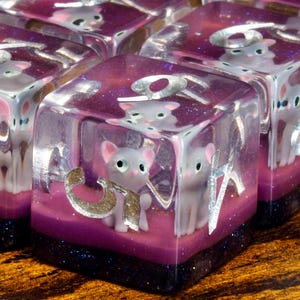 May include: Close-up of several dice with a cat theme. Each die is a clear cube with a silver number and a small grey cat figurine inside. The dice have a purple and glittery base.