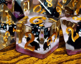 May include: A set of clear resin polyhedral dice featuring embedded miniature black and white cat figures. Each die has gold-colored numbers painted on the exterior surfaces. The dice have a translucent appearance with a light purple base tint.