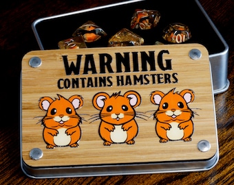 Puede incluir: Una caja de madera con tapa de metal y una etiqueta de advertencia que dice "WARNING CONTAINS HAMSTERS". La caja tiene tres hámsters de dibujos animados en la parte delantera, cada uno con una expresión diferente. La caja está abierta y hay seis dados transparentes dentro.