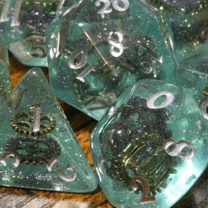 Op de afbeelding: Een set polyhedrale dobbelstenen gemaakt van doorschijnende lichtblauwe hars met ingebedde metalen tandwielonderdelen en fijne glitterdeeltjes. Elke dobbelsteen heeft scherpe randen en duidelijke, zilverkleurige cijfers die in het oppervlak zijn gegraveerd, wat een complex intern mechanisch ontwerp laat zien door het heldere materiaal.