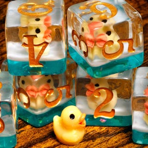May include: A set of clear resin dice with a teal blue base and a white layer, each containing a miniature yellow rubber duck. The dice feature gold-colored numbers on the exterior faces, and three loose yellow rubber ducks are placed nearby.