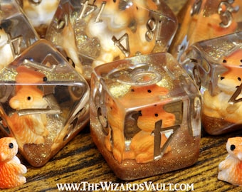 May include: A set of clear resin polyhedral dice containing miniature orange fox figurines and gold glitter, with metallic numbers. Two small orange fox figurines stand outside the dice. The text www.thewizardsvault.com is at the bottom.