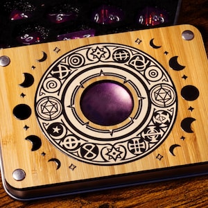 May include: A wooden dice box with a detailed, etched design featuring geometric shapes, moons, and stars. The box has a purple center and silver metal corners. Inside the open box, there are several purple dice. The box is on a wooden surface.