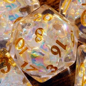 May include: A collection of polyhedral dice made from clear resin with an iridescent, shimmering internal texture. Each die features deeply engraved numbers painted in a metallic gold finish, including a twenty-sided die prominently displayed.