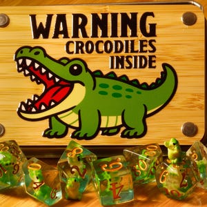 以下が含まれることがあります： 「WARNING CROCODILES INSIDE」の文字と漫画のアリゲーターのイラストが描かれた木製の看板。看板の前には、アリゲーターのフィギュアが入った緑と透明のサイコロがいくつかあります。