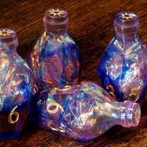 May include: Four iridescent glass bottles with gold numbers and starburst-shaped stoppers. The bottles are blue, purple, and gold, with a faceted design. One bottle is lying on its side.