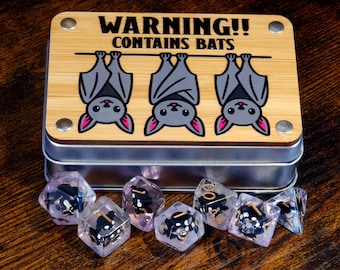 May include: A wooden box with the text "WARNING!! CONTAINS BATS" and three cartoon bats hanging upside down. The box sits on a metal tin, with several clear, purple-tinted dice with black numbers in front.