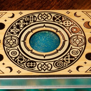 K&ouml;nnte beinhalten: Eine Holzkiste mit einem kreisf&ouml;rmigen Design, das verschiedene Symbole und eine blaue, glitzernde Mitte zeigt. Die Kiste hat einen Metallrahmen und vier Metallakzente.